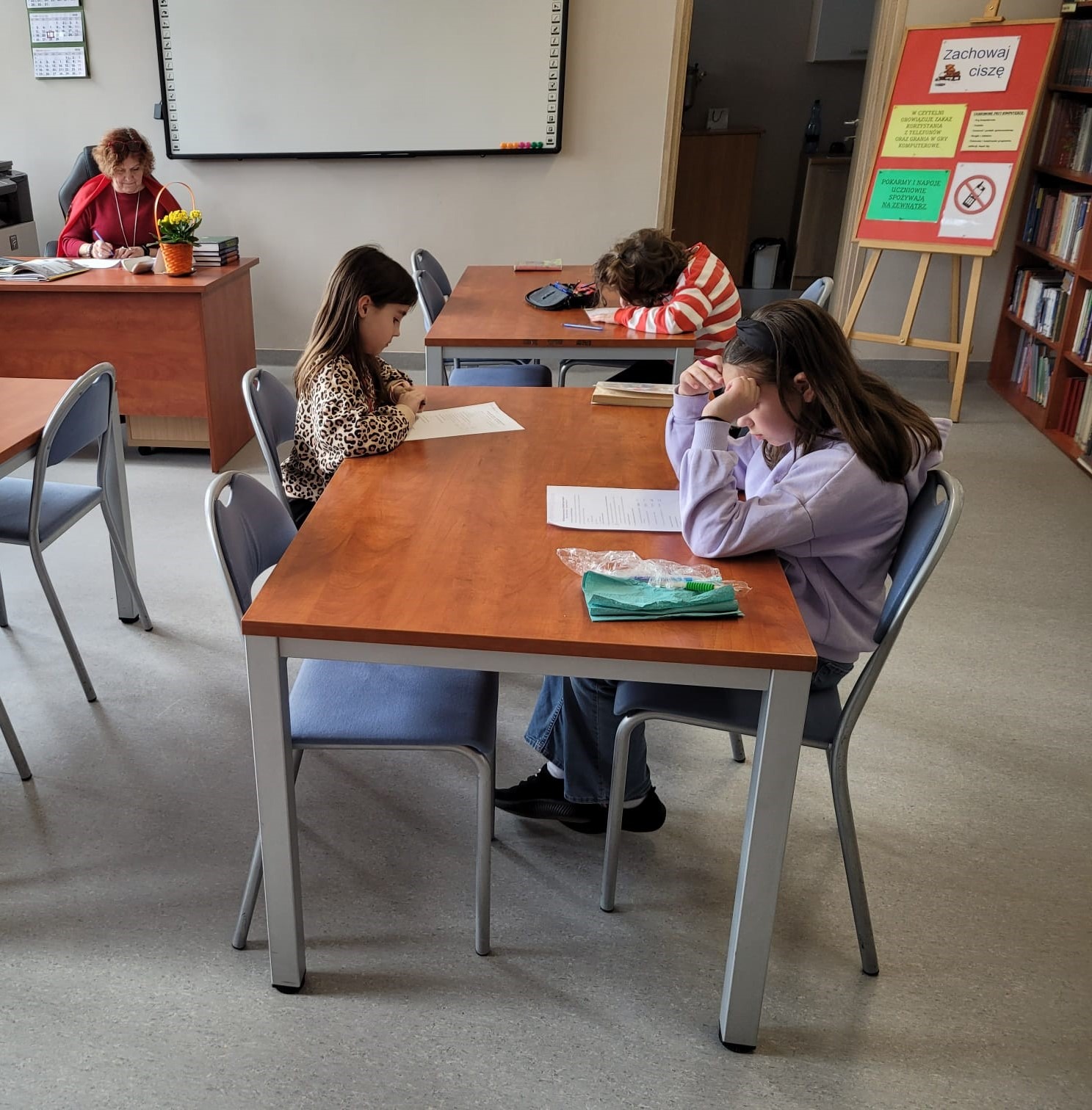 Na zdjęciu uczniowie w bibliotece siedzą w ławkach i piszą szkolne eliminacje konkursu. Za biurkiem siedzi pani Krystyna May