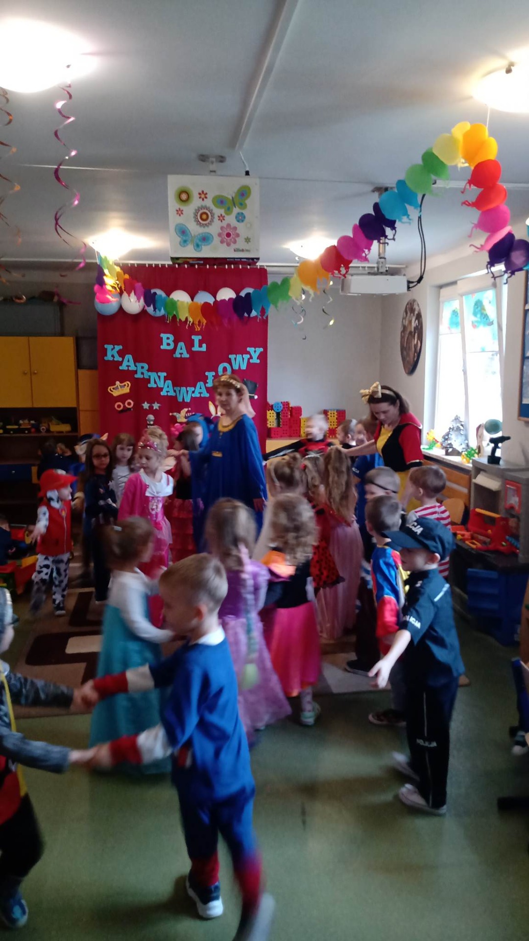 Dzieci i dwie nauczycielki w kolorowych strojach tańczą w rytm muzyki karnawałowej.