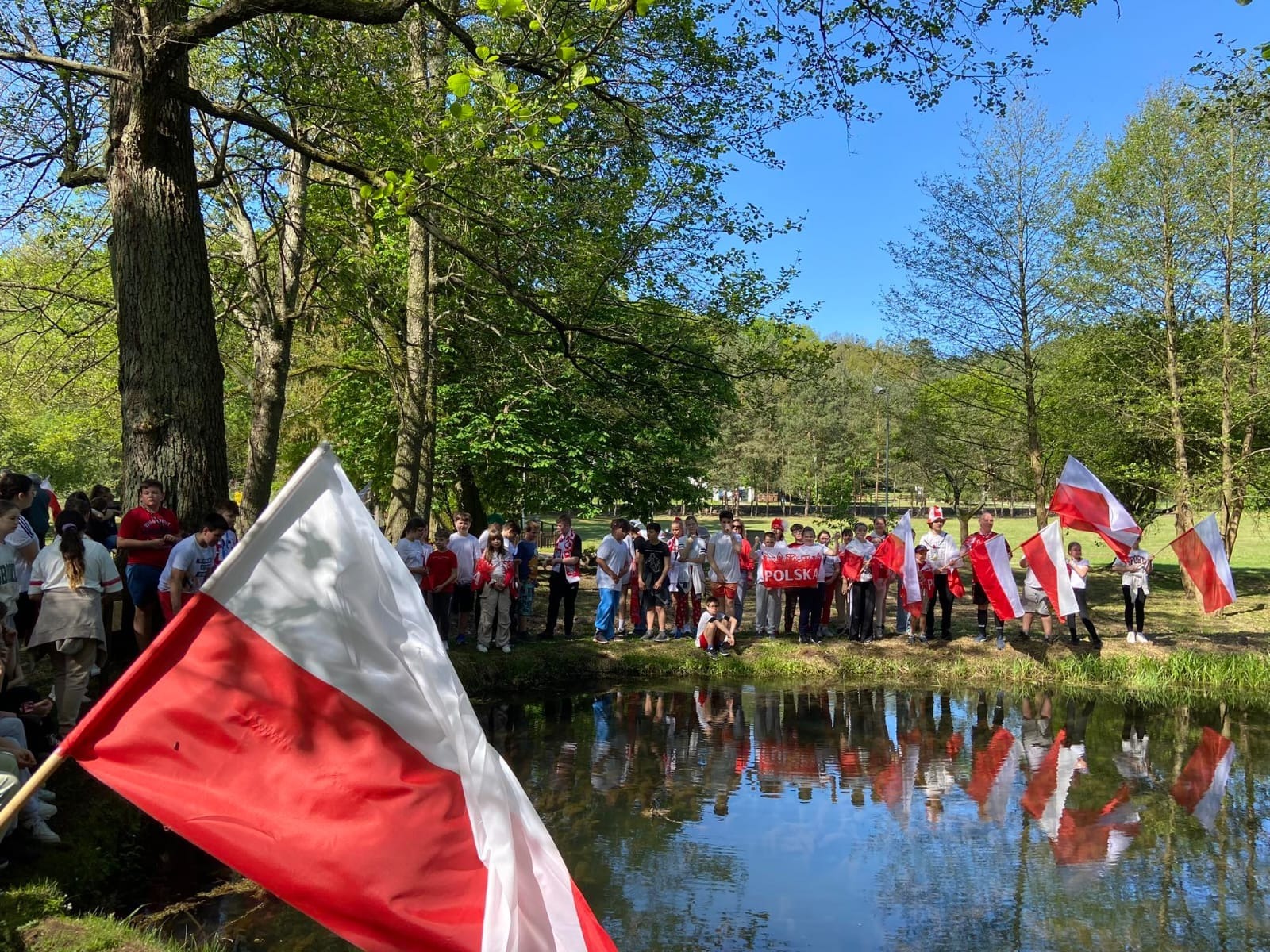Uczniowie w biało czerwonych strojach z flagami polski pozują przy stawie do zdjęcia.