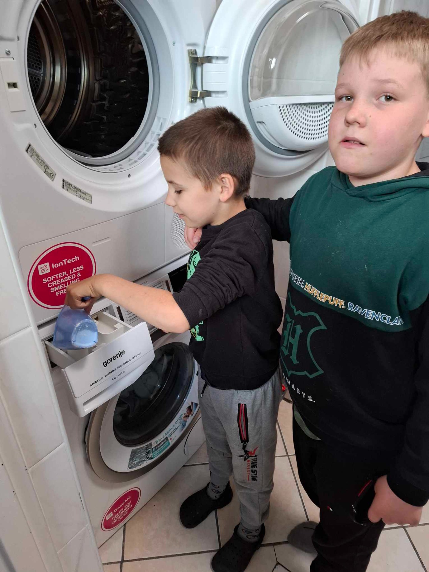 2 uczniów stoi obok pralki, jeden z nich wsypuje proszek do szufladki pralki