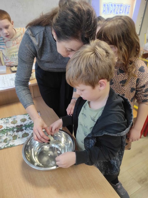 nauczycielka wraz z chłopcem leją wosk, przez klucz do miski z wodą, za nimi stoi dziewczynka, z oddali zza biurka zagląda chłopiec