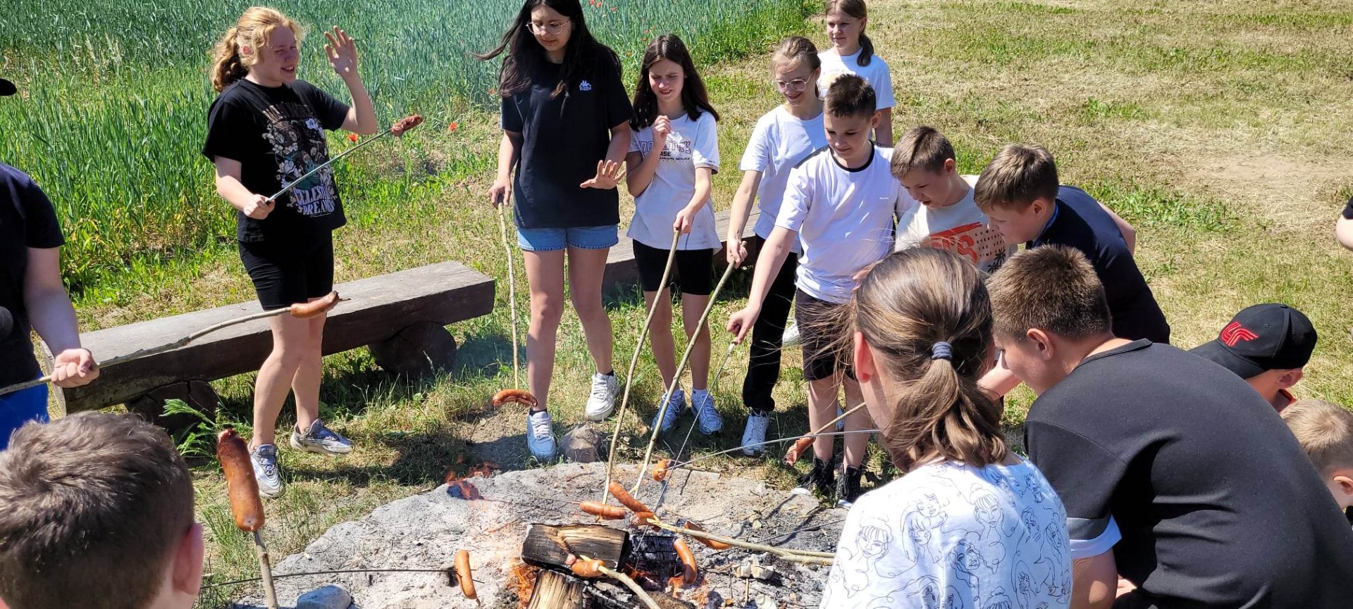 Uczniowie stoją przy ognisku, pieką kiełbaski.