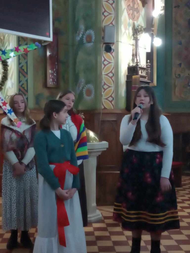 Zdjęcie przedstawia uczniów stojących w kościele przebranych za kolędników, Maryję, Józefa, aniołki, dzieci z Polski i Kolumbii biorących udział w przedstawieniu.