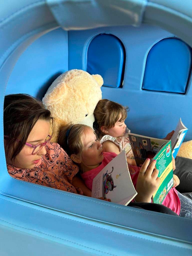 Trzy dziewczynki z klasy pierwszej b czyta książki w piankowym, niebieski domku. Jedna oparta jest o olbrzymiego misia.
