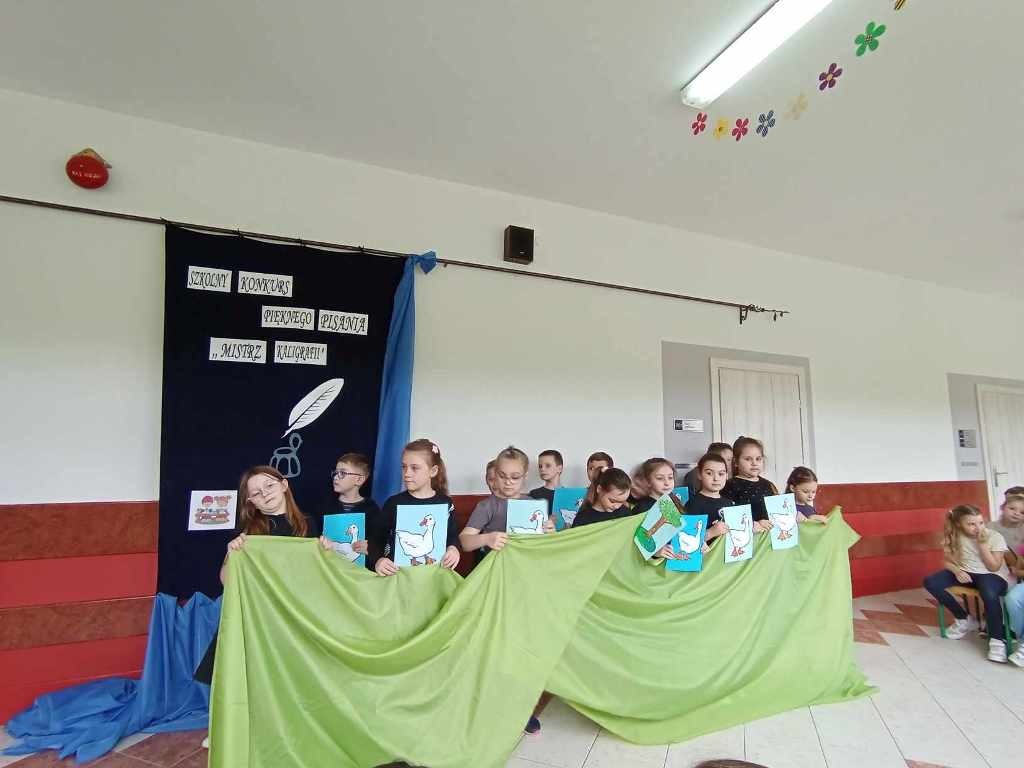 Zdjęcie przedstawia uczniów edukacji wczesnoszkolnej biorących udział w przedstawieniu. Uczniowie stoją za zielonym materiałem, a za nimi znajduje się dekoracja z napisem Szkolny Konkurs Pięknego Pisania Mistrz Kaligrafii 2024. W rękach trzymają niebieskie kartki z białymi gąskami.