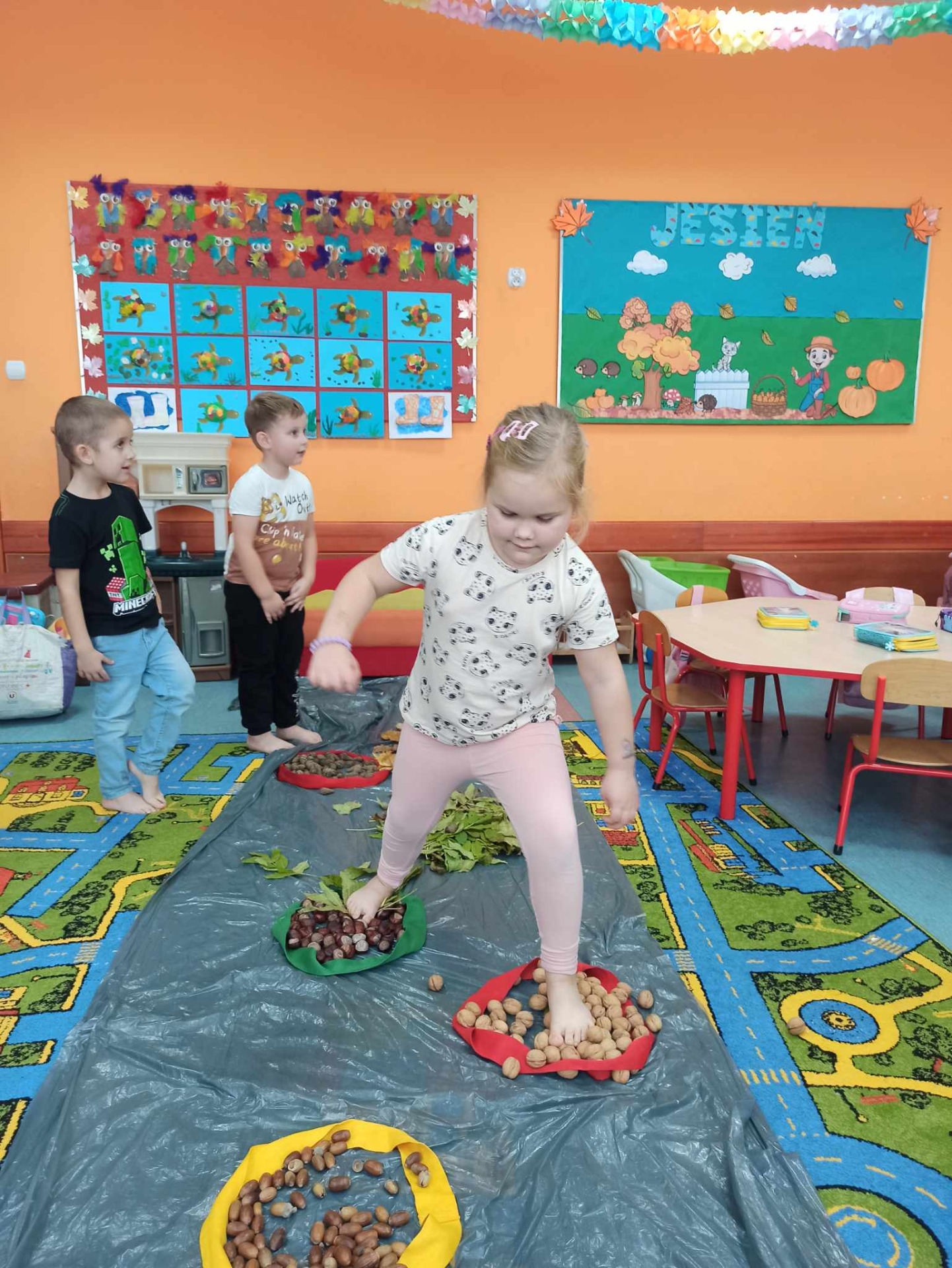 Dziecko spaceruje po torze sensorycznym, a grupa przedszkolaków siedzi na kolorowym dywanie obok toru sensorycznego