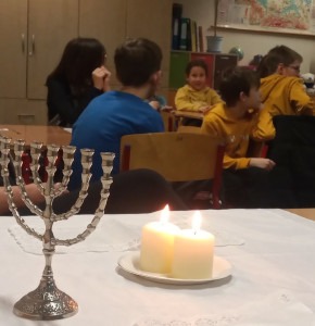 Na pierwszym planie menora i zapalone świeczki, w głębi klasy uczniowie przy stolikach