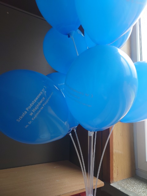 Pięć niebieskich balonów z logo szkoły umieszczonych w stojaku.
