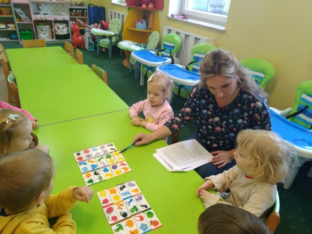 Opiekunka z dziećmi siedzi przy zielonym stoliku w sali . W ręku trzyma kartkę . Na zielonym blacie stolika leżą kolorowe kartki. Opiekunka wskazuje kredką wybrany element z ilustracji.