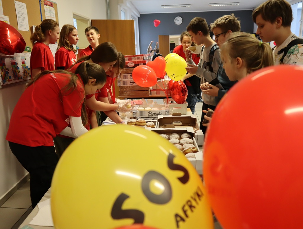 Uczniowie w czerwonych koszulkach z napisem "SOS dla Afryki" sprzedają pączki podczas przerwy szkolnej.
